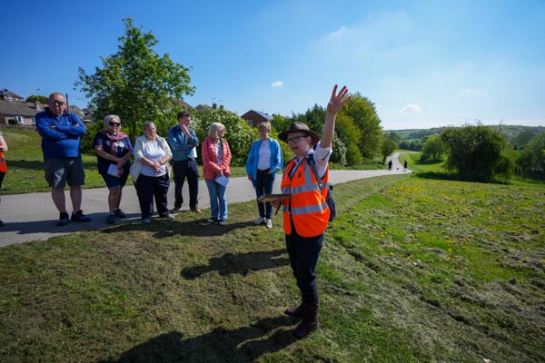 A large group gathered on the village green in sunshine while an organiser addresses them