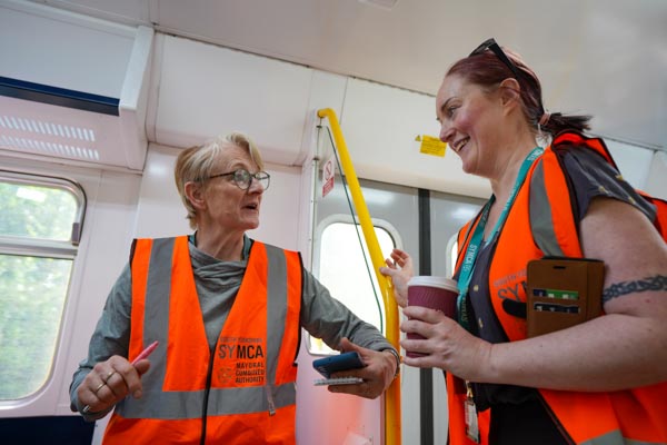 Two people in SYMCA hi-vis vests talking and laughing on board a Northern Trains service